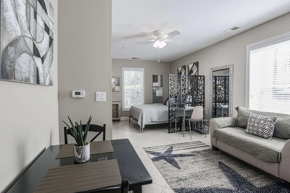 A comfortable living and dining space featuring a dining table set for two, a soft sofa with decorative pillows, and a bed in the background. The arrangement encourages both intimate meals and relaxed socializing, creating a flexible environment for entertainment or downtime.