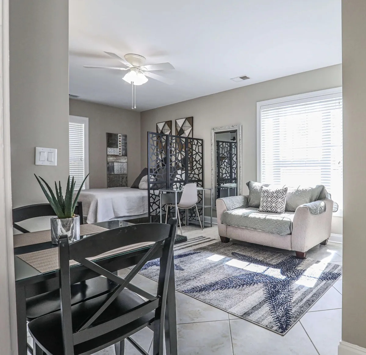 A stylish, sunlit studio featuring modern furnishings, a cozy sofa with plush cushions, a chic dining area perfect for shared meals, and a decorative room divider that adds both function and flair. Soft morning light streams through the window, illuminating the contemporary space and its textured area rug.