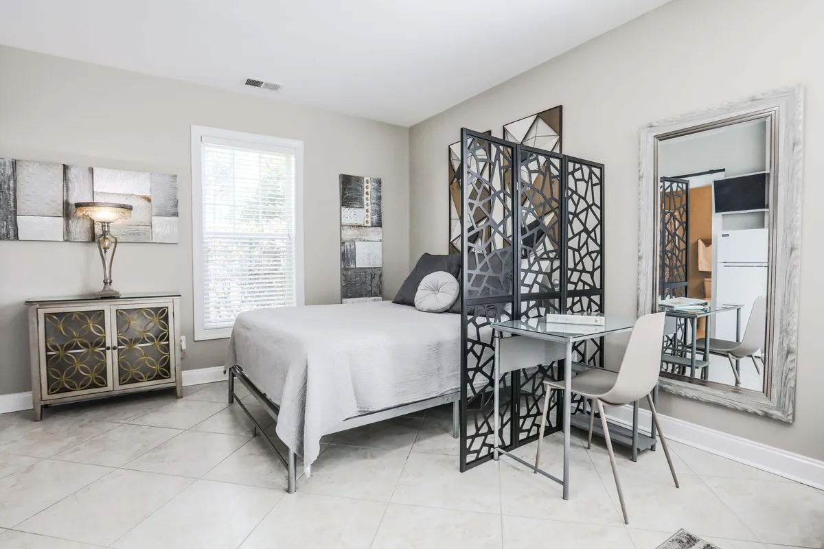 This tranquil interior shot focuses on the studio's sleeping area with a cozy bed, decorative screen, and a dedicated workspace featuring a desk and chair. Natural light filters in, emphasizing the calm atmosphere and modern décor. A mirror and lamp add both function and ambiance.