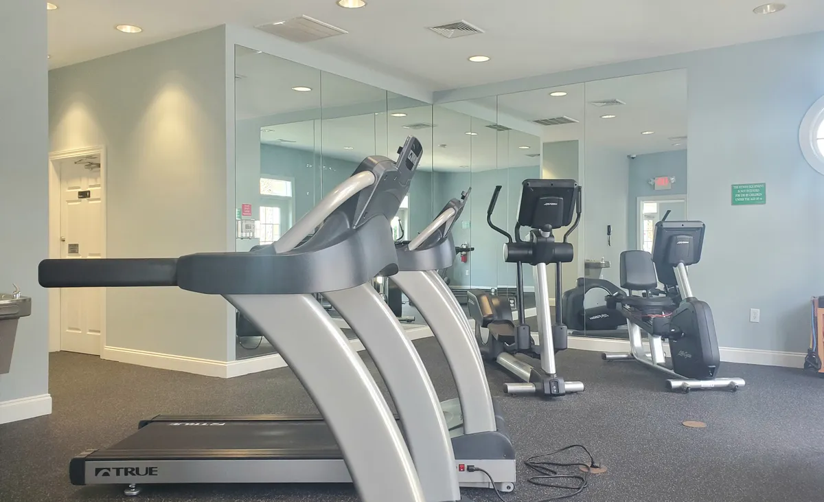 This interior image shows a bright, modern fitness center equipped with treadmill, elliptical machine, exercise bike, and mirrored walls. The space is designed to inspire both personal fitness goals and community workouts in a contemporary, motivational setting.