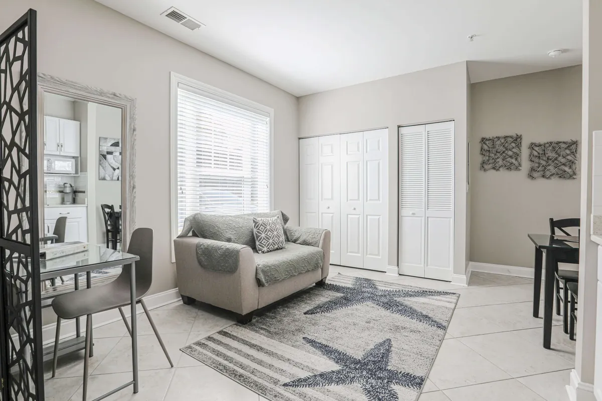 A welcoming living space with a plush sofa, a dining table set for intimate meals, a starfish-patterned rug, and a large mirror reflecting sunlight. Morning rays stream through sizable windows, filling the room with warmth and comfort.