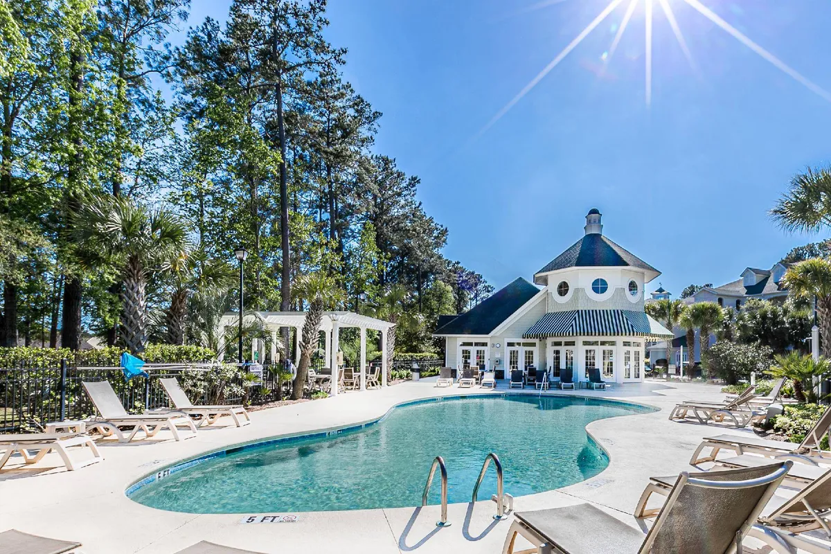 An inviting exterior view of the resort's swimming pool, surrounded by lounge chairs and lush greenery, with the clubhouse visible in the background. The space is designed for sunbathing, swimming, and socializing, capturing a lively and relaxing resort atmosphere.