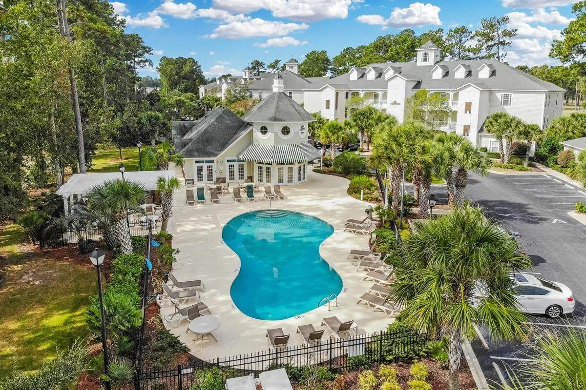 This exterior image features a sparkling swimming pool surrounded by palm trees, elegant lounge chairs, and the inviting clubhouse in the background. The area radiates resort-style luxury, with ample space for relaxation, sunbathing, and socializing.
