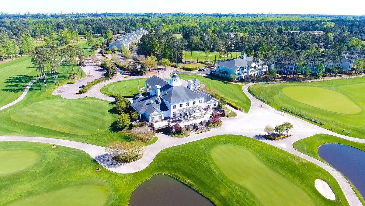 This exterior image features a sweeping view of the World Tour Golf Course with meticulously maintained greens, the elegant clubhouse, a tranquil pond, and mature trees bordering the landscape. The clubhouse, visible in the background, serves as a focal point, emphasizing the property's direct proximity to luxury golfing amenities and social spaces.